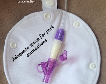 May include: A white circular fabric port cover featuring a black clip for attaching to clothing or a backpack, an extra snap for cord organization, and text stating "Clip for attaching to clothing or backpack," "Extra snap for cord organization," and "Adequate space for port connections."