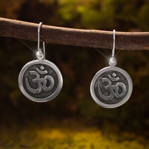 May include: A pair of silver-colored earrings with a circular pendant featuring the Om symbol. The earrings are suspended from a simple hook and are displayed against a blurred, natural background.