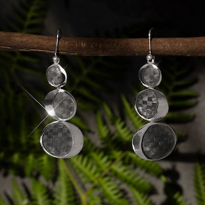 Puede incluir: Un par de pendientes plateados con un diseño de tres niveles. Cada nivel presenta un patrón texturizado y tejido dentro de una forma circular u ovalada. Los pendientes cuelgan de un gancho simple, mostrando una estética moderna y geométrica.