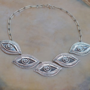 Puede incluir: Collar plateado con cinco colgantes con forma de ojo. Cada colgante presenta un diseño detallado de ojo con círculos concéntricos y un borde texturizado. El collar está conectado por una cadena y tiene un aspecto artesanal.