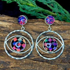 Può includere: Un paio di orecchini color argento. Ogni orecchino presenta una piccola pietra rotonda e iridescente nella parte superiore e un design più grande a forma di occhio nella parte inferiore. Il design a forma di occhio ha un motivo a scacchi colorato al centro. Gli orecchini sono su legno.
