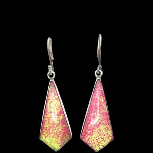 Può includere: Un paio di orecchini color argento con un design geometrico vibrante. Ogni orecchino presenta un pendente triangolare maculato rosa e giallo, incastonato in una cornice d'argento. Gli orecchini pendono da un semplice gancio.