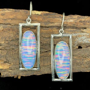 Può includere: Orecchini in argento con pietre di opale ovali e iridescenti incastonate in cornici rettangolari. Gli opali mostrano sfumature blu, rosa e gialle, in contrasto con l'argento. Gli orecchini hanno una chiusura a gancio. Gli opali misurano circa 2 cm di lunghezza.