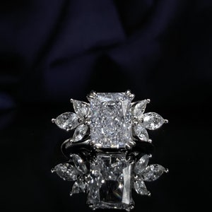 Pode incluir: Um anel de prata com uma grande pedra central de diamante retangular e transparente. O diamante é rodeado por diamantes menores e transparentes dispostos em forma de flor.