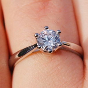 May include: A silver engagement ring with a round, clear diamond set in a six-prong setting.