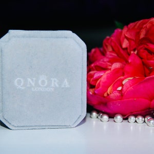 Pode incluir: Uma caixa de joias de veludo cinza com o texto "QNORA LONDON" impresso em branco.