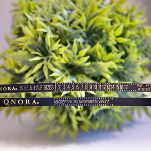 Pode incluir: Fita m&eacute;trica preta com letras douradas. A fita tem uma r&eacute;gua com os n&uacute;meros de 1 a 17 e o alfabeto. O texto na fita diz "QNORA. SIZE & HALF SIZES 1 2 3 4 5 6 7 8 9 10 11 12 13 14 15 16 17 PAT. PEND. 9101693".