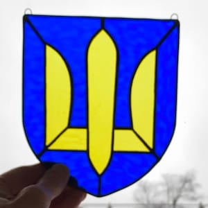 May include: A stained glass window depicting the Ukrainian coat of arms, a yellow trident on a blue shield.