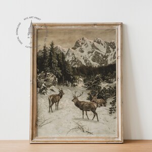 May include: A framed vintage print depicting a snowy mountain landscape with three deer in the foreground. The deer are brown and have antlers. The mountains are in the background and are covered in snow. The sky is a light blue colour.