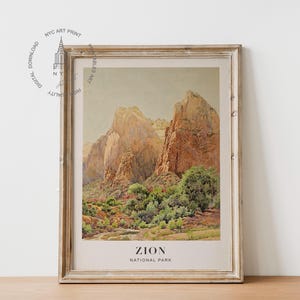 May include: Framed print of Zion National Park. The artwork depicts towering rock formations in shades of tan and brown, with green foliage at the base. The print is in a distressed, light-colored wooden frame. The text "ZION NATIONAL PARK" is at the bottom.