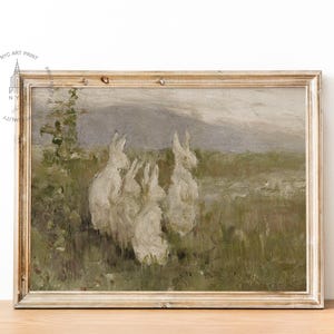 May include: A vintage painting of a group of white rabbits in a field. The rabbits are gathered together in a grassy area with a mountain range in the background. The painting is framed in a light wood frame.