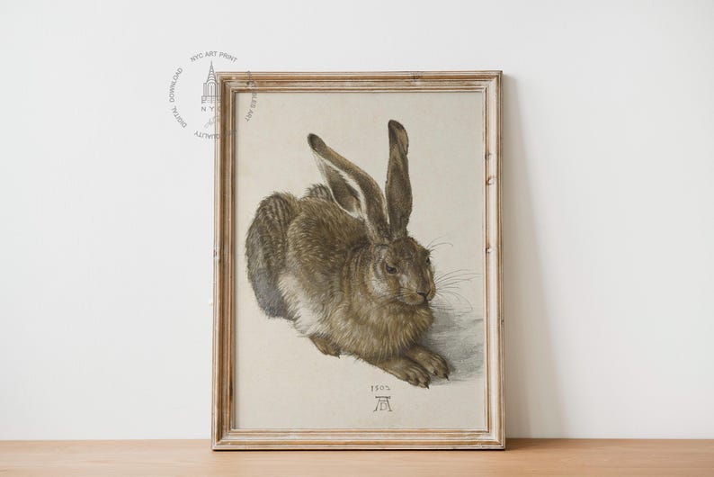 May include: A framed print of a brown hare, depicted in a realistic style. The hare is lying down with its head turned to the side. The print is in a light brown wooden frame and has the year 1502 and the letter A printed in the bottom center.