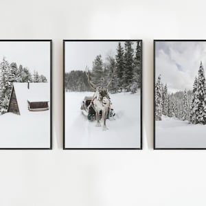 May include: Set of three framed winter landscape prints. The prints feature snowy scenes: a cabin, a reindeer pulling a sled, and a forest of snow-covered trees. The prints are in black frames.
