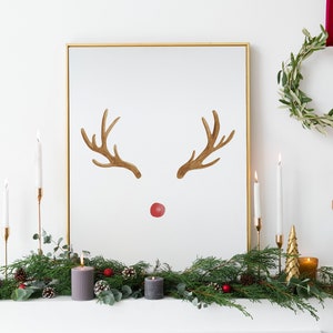 May include: A gold framed picture with a watercolor reindeer illustration. The reindeer has brown antlers and a red nose. The picture is on a white mantel with a green garland, candles, and a small gold Christmas tree.