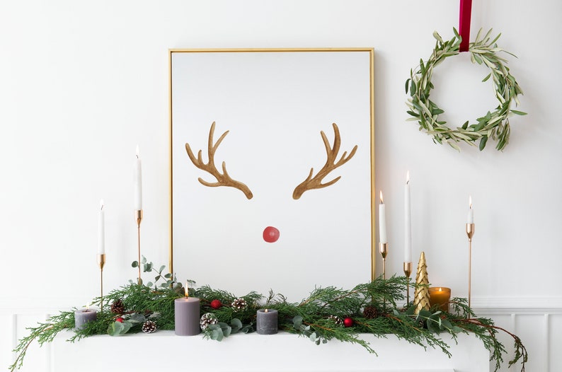 May include: A gold framed picture with a watercolor reindeer illustration. The reindeer has brown antlers and a red nose. The picture is on a white mantel with a green garland, candles, and a small gold Christmas tree.