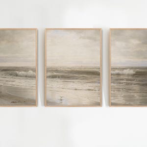 May include: Three framed art prints depicting a seascape with a beach, waves, and cloudy sky. The prints have a vintage aesthetic with muted tones of beige, gray, and brown. The frames are a light wood color. The prints are labeled "NYC ART PRINT".