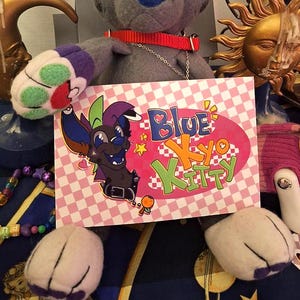 May include: A plush toy dog with blue and gray fur, wearing a red collar. A sign with the text "Blue Kyo Kitty" is in front of the toy. The background includes decorative items like a sun sculpture and a candle holder.