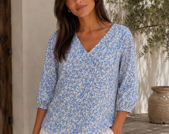 May include: A light blue V-neck blouse featuring a dense pattern of small white and yellow flowers. The top has three-quarter length sleeves with gathered cuffs and a straight hemline, crafted from a lightweight fabric with a soft texture.
