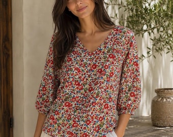 May include: A V-neck blouse featuring a dense floral pattern with red, blue, green, and yellow blossoms on a white background. The top has three-quarter length sleeves with gathered elastic cuffs and a relaxed fit, paired with light-colored trousers.