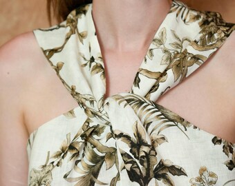 May include: A sleeveless top featuring a cream-colored base with a dark brown botanical print. The garment has a high neckline with a central twisted knot detail that creates a draped effect across the chest and shoulders, showcasing a tropical foliage pattern.