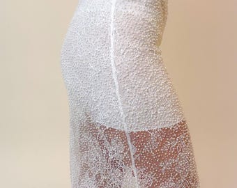 May include: A white midi skirt featuring an intricate floral lace overlay embellished with numerous small white beads. The skirt has a high-waisted fit with a solid white lining that ends above the knee, while the lace hem falls below the knee.