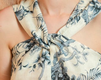 May include: A sleeveless top featuring a cream-colored base with a blue botanical print depicting monkeys and tropical foliage. The garment has a distinctive knotted detail at the center of the neckline, creating a draped effect across the chest.