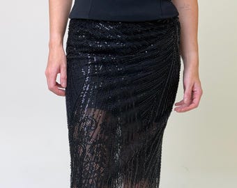May include: A black sleeveless top paired with a black midi skirt featuring intricate beadwork, sequins, and lace detailing. The skirt has a wrap-style silhouette with a sheer hemline and dense vertical beaded patterns throughout the fabric.