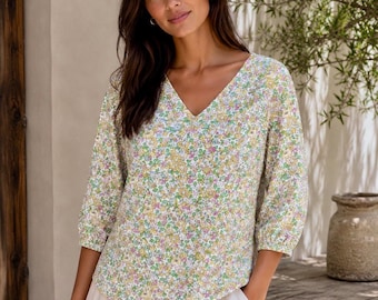 May include: A V-neck blouse featuring a small-scale floral pattern in shades of yellow, green, and purple on a white background. The top has three-quarter length sleeves with gathered cuffs and a straight hemline, paired with light-colored trousers.
