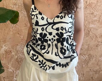 May include: A sleeveless camisole top featuring a white base with a bold black floral pattern, a V-neckline, and thin black shoulder straps, paired with plain white shorts. The top has a distinct horizontal black border design near the hem.
