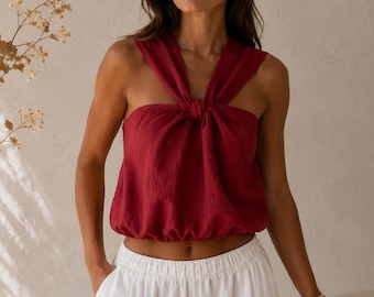 May include: A sleeveless deep red top featuring a knotted halter neckline and a gathered, elasticized hemline, paired with white shorts that have an elastic waistband and side pockets. The top is made of a textured fabric with a relaxed fit.
