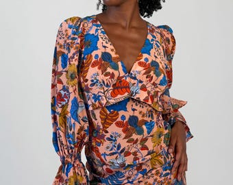 May include: A long-sleeved dress in a peach-colored fabric featuring a vibrant, multi-colored botanical print with blue, red, and yellow floral and leaf motifs. The dress has a V-neckline, puffed shoulders, and sleeves with gathered, ruffled cuffs.