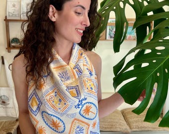 May include: A sleeveless halter top featuring a cream-colored fabric with a repeating pattern of blue and orange illustrations, including palm trees, suns, and geometric shapes. The top has a draped neckline and a lightweight, textured appearance.