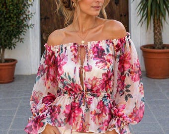 May include: Off-the-shoulder floral blouse with long, sheer sleeves. The blouse features a white base with a vibrant pink floral pattern. It has a drawstring neckline and a peplum waist, creating a feminine silhouette. The model is wearing cream-colored pants.
