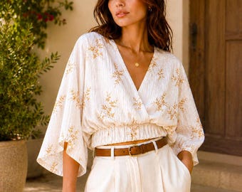 May include: Cream-colored wrap top with a floral pattern and wide sleeves. The top is paired with white pants and a brown belt. The outfit is completed with a brown handbag. The style is casual and elegant.