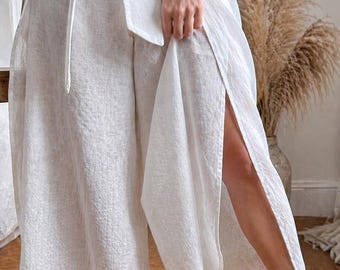 May include: White linen palazzo pants with a high waist and a matching wrap-around belt. The pants have a wide leg with a high side slit. The model is wearing patterned heeled sandals. The outfit is a neutral color.