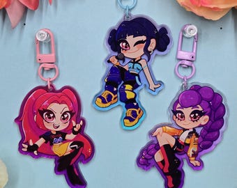 May include: Three cartoon character keychains with pink, blue, and purple hair. Each keychain features a different character design with a silver clasp. The characters are wearing colorful outfits and are set against a light blue background.