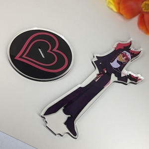 Puede incluir: Un soporte de acr&iacute;lico negro con un dise&ntilde;o de coraz&oacute;n rojo y una figura de acr&iacute;lico transparente de un personaje de dibujos animados con cuernos rojos, un atuendo blanco y negro y una m&aacute;scara amarilla y roja. El personaje sostiene un cuchillo rojo.