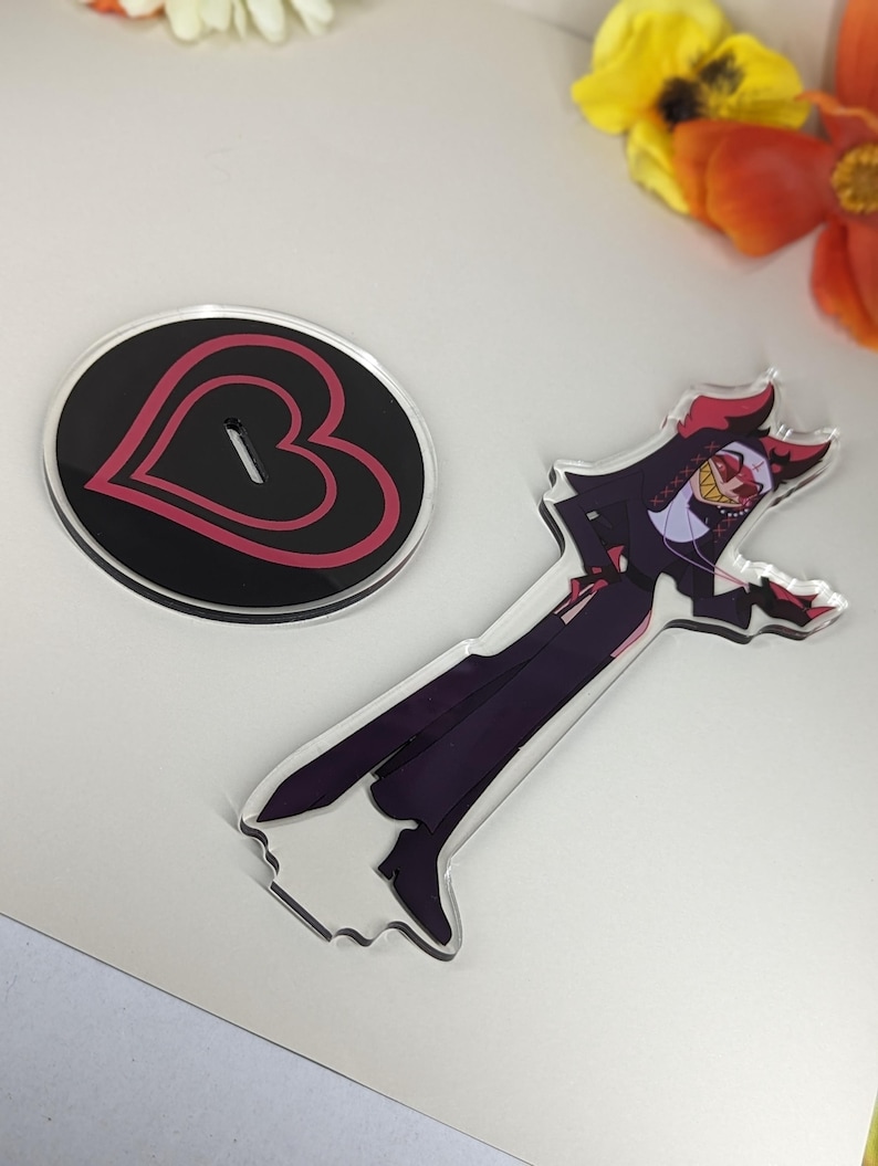 Puede incluir: Un soporte de acr&iacute;lico negro con un dise&ntilde;o de coraz&oacute;n rojo y una figura de acr&iacute;lico transparente de un personaje de dibujos animados con cuernos rojos, un atuendo blanco y negro y una m&aacute;scara amarilla y roja. El personaje sostiene un cuchillo rojo.