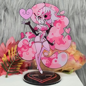 May include: A pink acrylic standee featuring a cartoon character with pink hair and a matching outfit. The character is holding a heart-shaped object and has a heart-shaped design on the stand. The background is blurred with pink and white accents.
