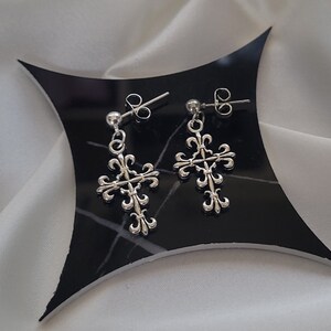 May include: A pair of silver cross earrings with a fleur-de-lis design. The earrings are on a black background.