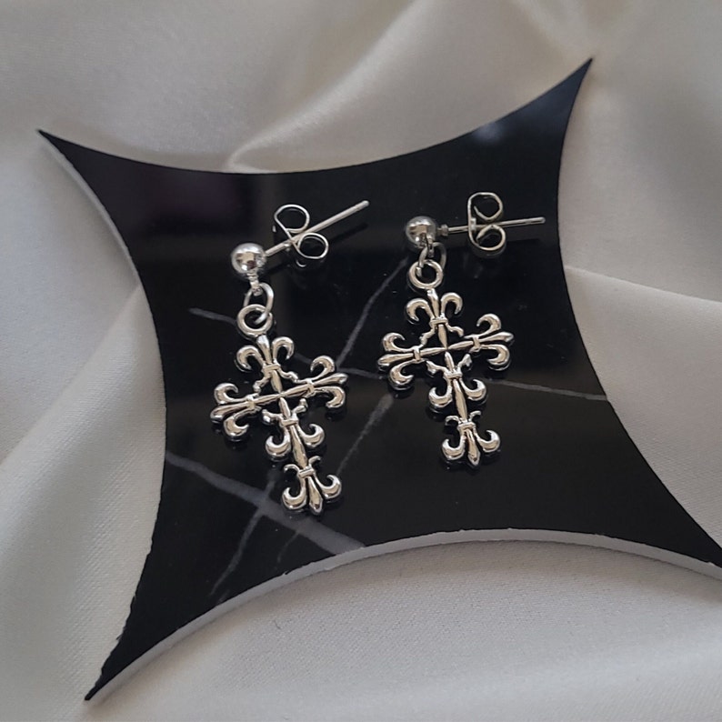 May include: A pair of silver cross earrings with a fleur-de-lis design. The earrings are on a black background.