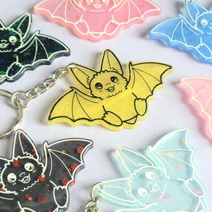 May include: A collection of bat-shaped keychains in various colors including yellow, black with red glitter, blue, and pink. Each keychain features a cartoon bat with outstretched wings, pointed ears, and white line details defining the face and wings.