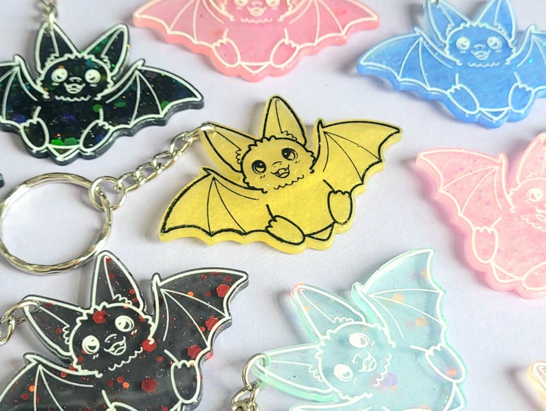 May include: A collection of bat-shaped keychains in various colors including yellow, black with red glitter, blue, and pink. Each keychain features a cartoon bat with outstretched wings, pointed ears, and white line details defining the face and wings.