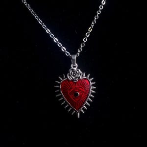May include: A silver chain necklace with a red heart pendant. The heart has a silver eye design and is surrounded by silver spikes.