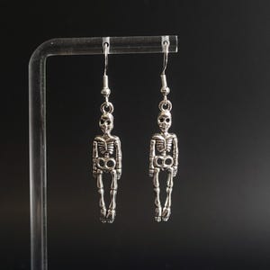 May include: A pair of silver-colored skeleton earrings. Each earring features a detailed miniature skeleton pendant hanging from a hook. The skeletons have articulated arms and legs, and are displayed against a dark background.