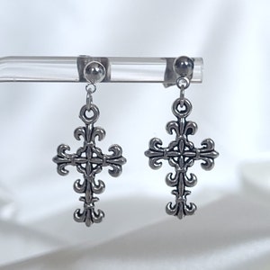 May include: A pair of silver cross earrings with a fleur-de-lis design. The earrings are on a clear display stand.