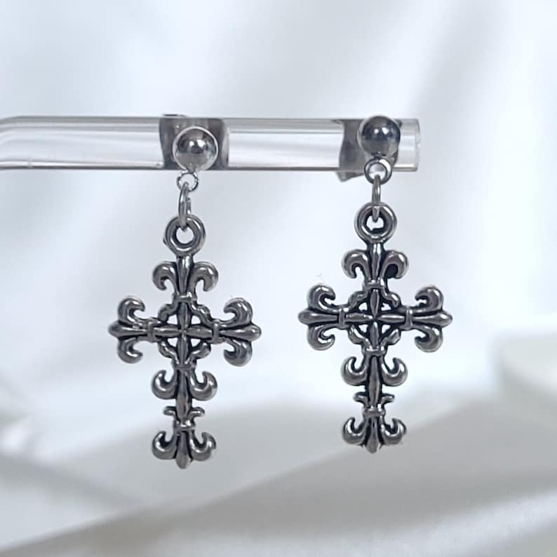 May include: A pair of silver cross earrings with a fleur-de-lis design. The earrings are on a clear display stand.