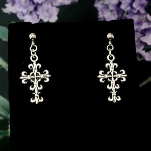 May include: A pair of silver cross earrings with a fleur-de-lis design. The earrings are hanging from a black background.
