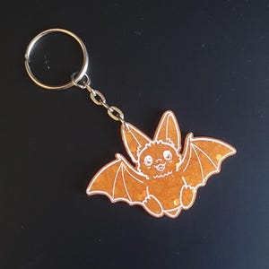 May include: A keychain featuring an orange glitter resin bat charm with white line art detailing the bat's face, ears, and wings. The charm is attached to a silver-toned metal split ring by a short chain, creating a decorative accessory.