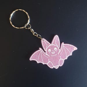 May include: A pink glittery bat keychain featuring a cartoon-style bat with large ears and outstretched wings. The bat has white line art details and is attached to a silver-colored metal split ring by a short chain link, creating a decorative accessory.